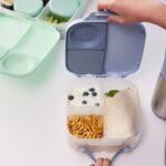 Manos colocando la comida en los compartimentos de la fiambrera infantil b.box