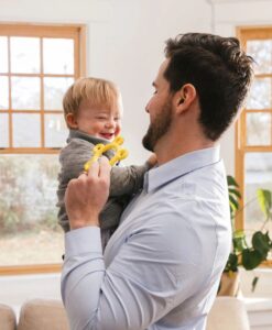Padre sonriente sujetando a su bebé, que muerde el mordedor Donut amarillo de ARK junto a una ventana