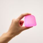 Gum Droppable de J'adore: mano sujetando una gominola rosa entre el pulgar y el índice