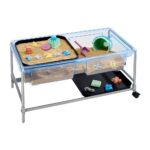 Mesa sensorial de agua y arena Edx: bandeja con arena y juguetes sobre el soporte y la otra tapa en la parte baja