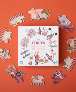 Puzzle redondo Circus round de Londji: caja cuadrada del circo rodeada de piezas sobre fondo naranja