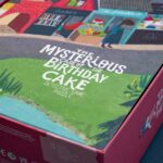 Puzzle de misterio de Londji: esquina de la caja con el título sobre el pueblo ilustrado